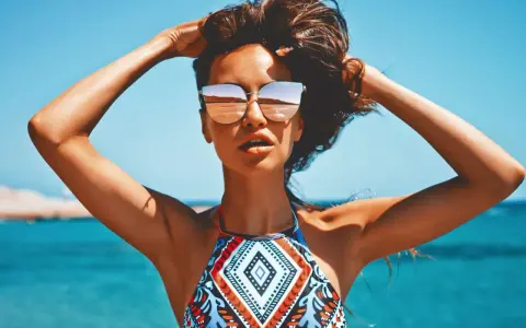 Kobieta w okularach przeciwsłonecznych na plaży, z rękami w gorze. Jej skóra ma piękny, złocisty odcień, jakby świadczył o wspaniałych beta karoten efektach.