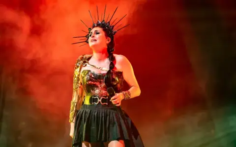 Wokalistka Within Temptation w koronie i błyszczącej kreacji, z mikrofonem w dłoni, na tle czerwonej mgły.
