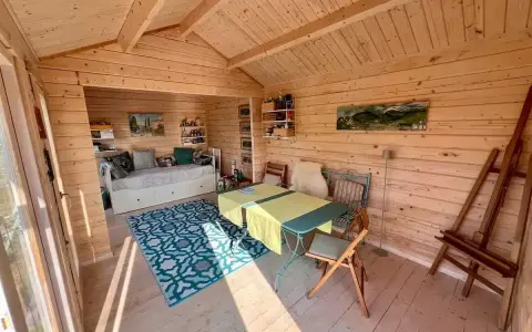 Gemütlicher Gartenhaus Innenausbau mit Tagesbett, Esstisch und Staffelei. Holzwände und Decke schaffen eine warme Atmosphäre.