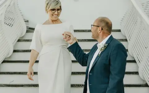 Elegancka kobieta po pięćdziesiątce w sukience do ślubu cywilnego, trzymająca za rękę pana młodego w granatowym garniturze.