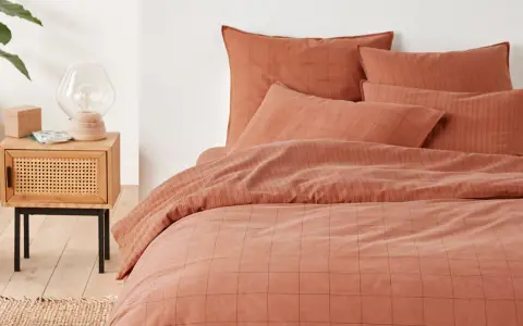 Cama con funda nordica terracota a cuadros y rayas. Mesita de noche de madera con lámpara y planta.