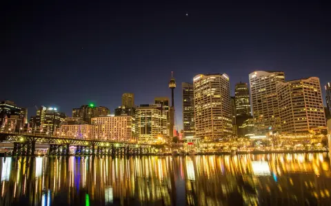 Sydney nocą, z mostem i odbijającymi się w wodzie światłami. Zastanawiasz się, ile się leci do Australii, patrząc na ten widok?
