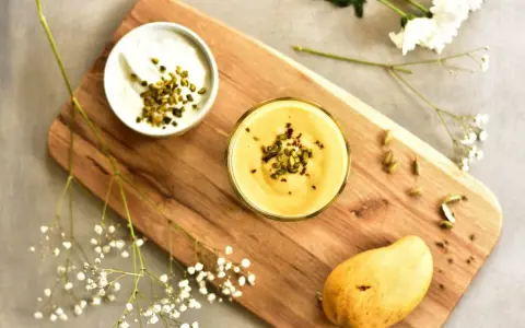 Aksamitny mango lassi w szklance, posypany pistacjami. Obok miseczka jogurtu, dojrzałe mango i kardamon.