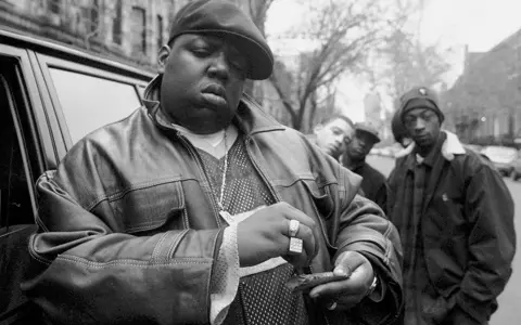 The notorious B.I.G. jak zginął. Raper w skórzanej kurtce i czapce, z towarzyszami, przed samochodem.