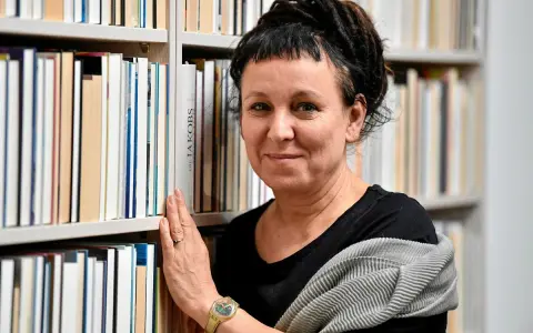 Olga Tokarczuk, w swoim wieku, otoczona książkami, z dłonią na grzbietach.