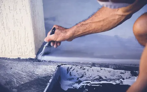Mężczyzna maluje pędzlem szarą farbą betonową przy ścianie. Widać kuwetę z farbą.