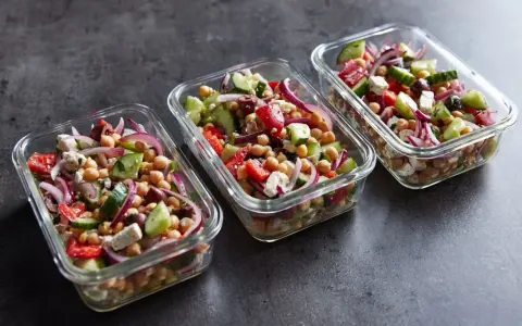 Trzy szklane pojemniki wypełnione kolorową sałatką z ciecierzycą, ogórkiem, pomidorem, czerwoną cebulą i fetą. Idealna na lunch.