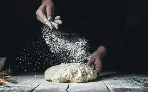Händer strör mjöl över en deg. Det är en bild av brödbakning med svenskt mjöl.