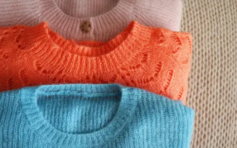 Gestapelte Pullover in Rosa, Orange und Blau. So kannst du deine **Wolle richtig waschen** und sie bleibt schön.