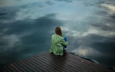 Eine Frau sitzt allein auf einem Steg, blickt aufs Wasser. Die Spiegelung der Wolken im Wasser verstärkt das Gefühl der **Angst vor dem Alleinsein**.