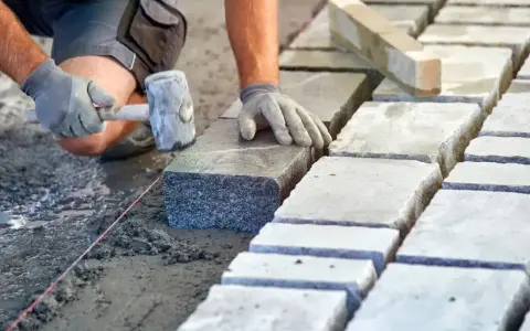Ręce w rękawiczkach układają kamienne kostki na podsypce. Widać młotek i sznur geodezyjny. Prawidłowe suchy beton proporcje to klucz do trwałej nawierzchni.