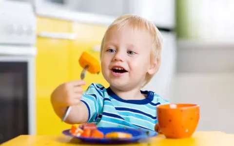 Niño rubio con tenedor y zanahoria, aprendiendo a comer solo. ¡Ayudame a hacerlo por mi mismo!