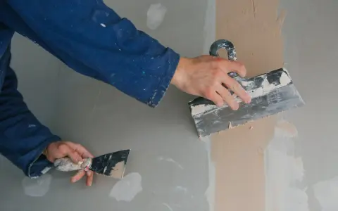 Ręce w niebieskim kombinezonie nakładają gładź szpachlową na ścianę za pomocą kielni.