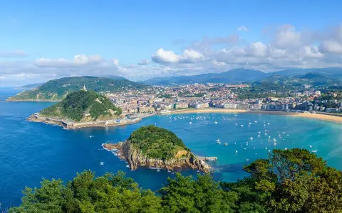 Widok na zatokę La Concha w Donostia San Sebastian, z zielonymi wzgórzami, miastem i licznymi łodziami na turkusowej wodzie.