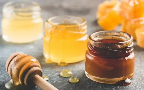 Trois pots de miel aux teintes variées, du clair au foncé, et une cuillère en bois dégoulinante. Quel miel pour pain d'épices ? Le choix est là !