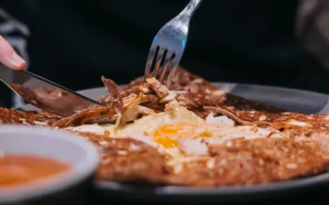 Une galette garnie d'œuf et de poulet, un délice des spécialités de Bretagne, est coupée avec un couteau et une fourchette.
