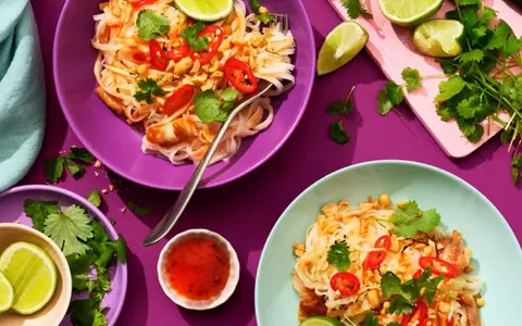 Två skålar med kyckling pad thai, toppad med chili, koriander och jordnötter. Limeklyftor och en liten skål med chilisås finns bredvid.