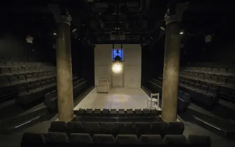 Pusta scena w **teatrze stu** miejsc, z rzędami siedzeń po bokach. Na scenie łóżko, białe krzesło i reflektory.