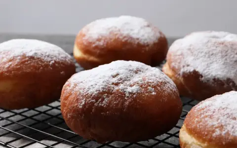 Pięć puszystych, pieczonych paczków, oprószonych cukrem pudrem, leży na czarnej kratce.