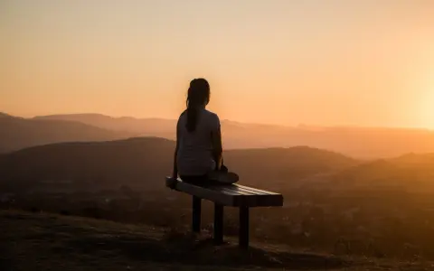 Eine Person sitzt auf einer Bank und blickt in die Ferne. Die untergehende Sonne taucht die Hügellandschaft in warmes Licht, ein Moment, um im Reinen zu sein.