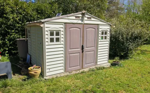 Un abri de jardin en plastique beige, avec des fenêtres et une double porte, est prêt pour son aménagement. Un petit panneau solaire est installé sur le toit.