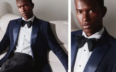 A man in a dark blue dinner jacket vs tuxedo, with a satin lapel and black bow tie, poses on a textured sofa.