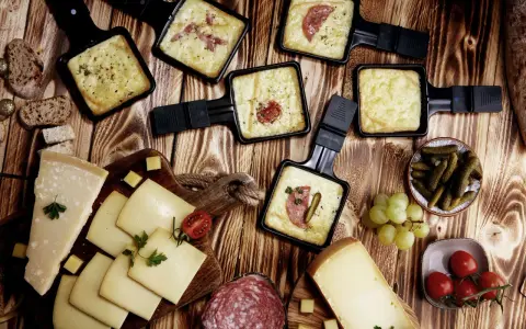 Raclette-Pfännchen mit geschmolzenem Käse, dazu Brot, Käse, Salami, Cornichons und Trauben. Perfekt für einen gemütlichen Abend mit **Wein zum Raclette**.