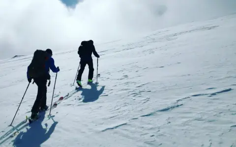 Två personer vandra på vintern uppför en snöklädd sluttning med skidutrustning och ryggsäckar.