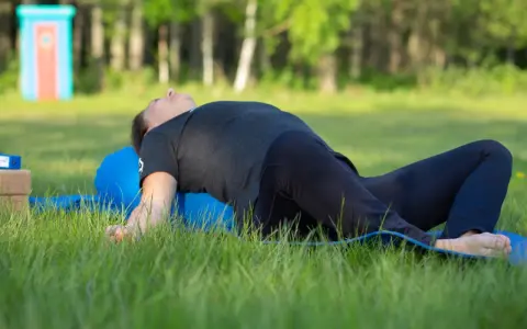 Person ligger på en blå yogarulle i gräset, med böjda knän och armarna utåt. En lugn stund med yin yoga utomhus.
