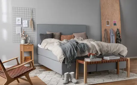 Ett mysigt sovrum grått med en stor säng, en trästol och en sittbänk. En rutig väggdekoration med bilder och en vas med bomull.