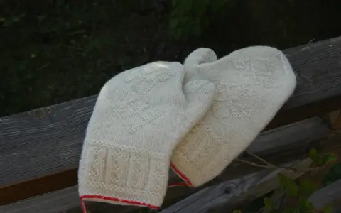 Två par vantar i naturvit ull, stickade med tvåändsstickning och fina mönster. De har en röd kant nertill.