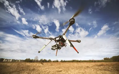 A drone hovers in a blue, cloudy sky over a dry, grassy field, hinting at the need to understand **Montana drone laws** before flying.