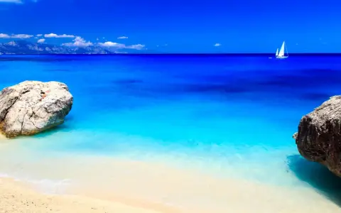 Spiaggia con rocce e mare cristallino, un veliero solca le acque turchesi. Potrebbe essere il mare più bello in Italia.