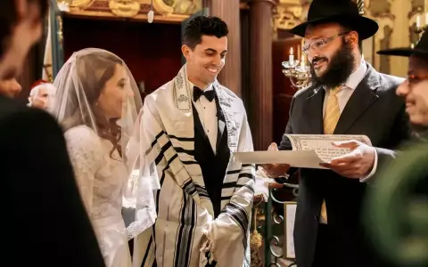 Novios sonríen durante su **matrimonio judío**. El novio lleva un talit y el rabino lee un documento.