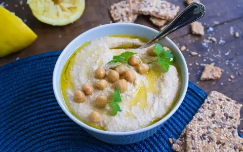 Cremiger Hummus, garniert mit Kichererbsen und Petersilie, daneben Zitronenspalten und knusprige Cracker. Das ist das beste Hummus Rezept!
