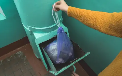 Ręka w żółtym swetrze wrzuca worek ze śmieciami do zsypu w bloku.