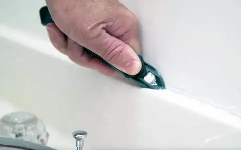 A hand uses a tool to smooth fresh caulk along a bathtub seam, demonstrating how to caulk for a clean finish.