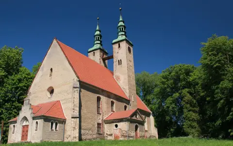 Stary kościół w stolcu z dwiema wieżami i zielonymi dachami, otoczony bujną zielenią drzew pod błękitnym niebem.