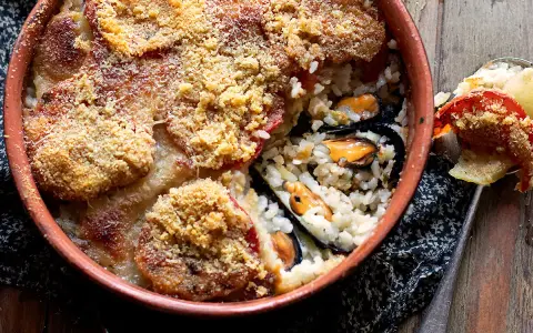 Piatto invitante di riso patate e cozze, gratinato con pomodoro e pangrattato, servito in una terrina di terracotta.