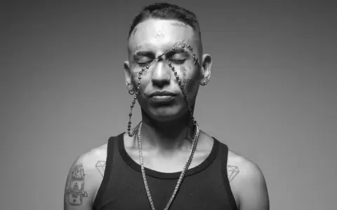 Un joven con tatuajes en la cara y el cuerpo, luciendo un collar de cuentas y una cadena de diamantes. Su estilo neo pistea barcelona es inconfundible.