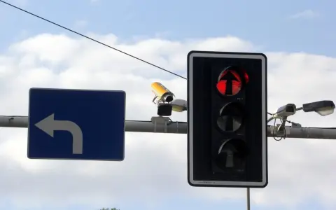 Rejestrator przejazdu na czerwonym świetle, widoczny na tle nieba, wraz z sygnalizacją świetlną i znakiem nakazu skrętu w lewo.