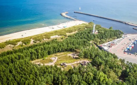 Widok z lotu ptaka na wybrzeże z piaszczystą plażą, lasem i portem. Wśród drzew widoczne są historyczne bunkry nad morzem.