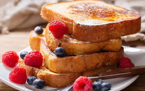 Una pila de tostadas francesas doradas, decoradas con frambuesas y arándanos, perfectas para aprender cómo se hacen las tostadas francesas.