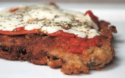 En närbild på en gyllene milanesa napolitana med smält ost, tomatsås och örter.