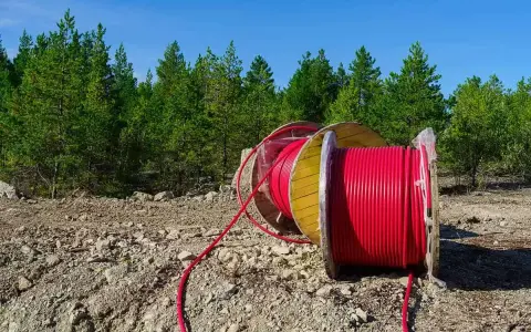 Czerwony kabel na drewnianym bębnie, przygotowany do uzbrojenia działki. W tle las i błękitne niebo.