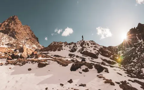 Ein Wanderer steht auf dem Gipfel des Jbel Toubkal, umgeben von schneebedeckten Felsen und einem strahlenden Sonnenschein.