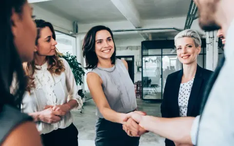 Uśmiechnięte kobiety w biurze, jedna z nich ściska dłoń partnerowi, symbolizując udaną współpracę w spółce partnerskiej.