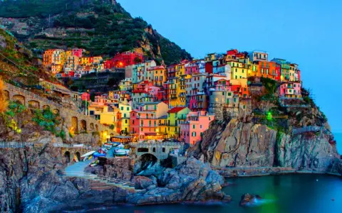 Case colorate aggrappate alla scogliera, un incantevole scorcio di quali sono le 5 Terre, con barche ormeggiate e il mare blu.