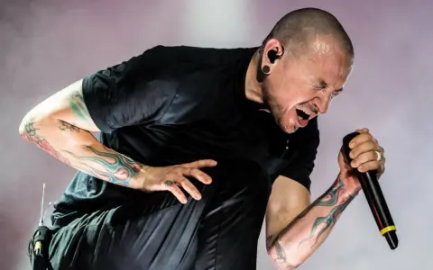 Chester Bennington z Linkin Park, z tatuażami na ramionach, śpiewa do mikrofonu z pasją.
