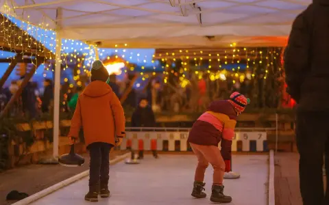Kinder spielen Eisstockschießen auf einem Weihnachtsmarkt in Berlin. Lichterketten leuchten im Abendlicht.